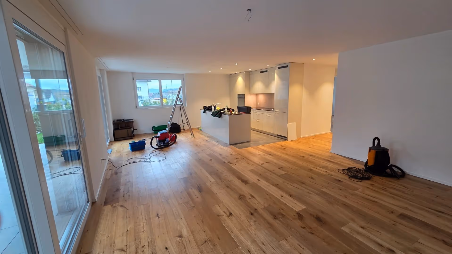 Offenes, helles Wohnzimmer mit Holzparkettboden und eingebauter moderner Küche, Werkzeuge und Leiter darauf verteilt.