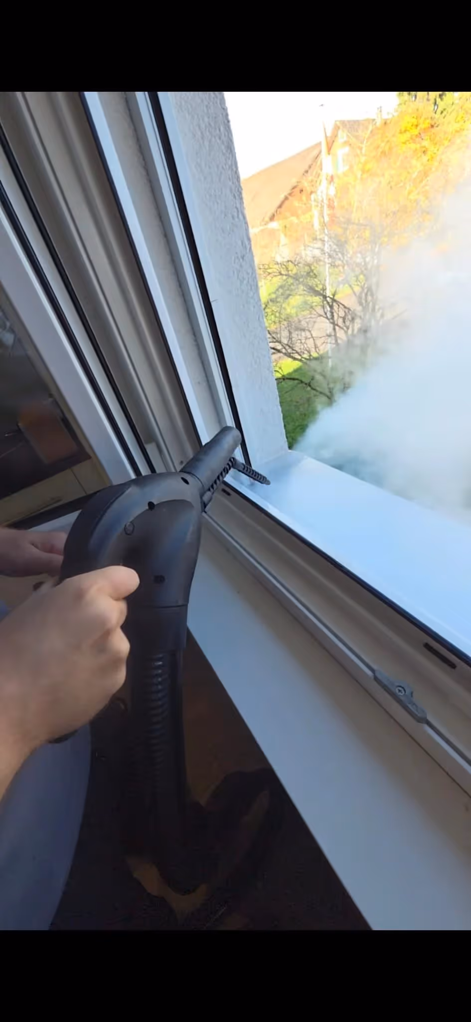 Person benutzt einen Dampfreiniger, um ein Fenster von innen zu säubern, wobei Dampf aus der Düse strömt.