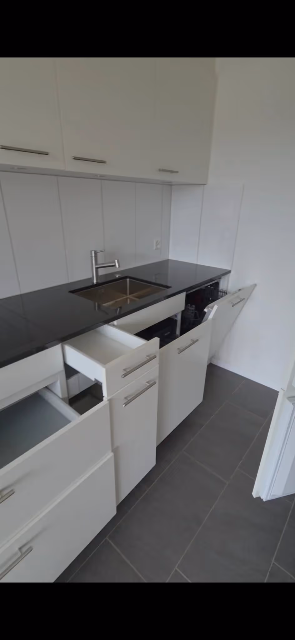 Moderne weiße Küche mit schwarzer Arbeitsplatte, Edelstahlspüle und geöffneten Schubladen und Geschirrspüler.